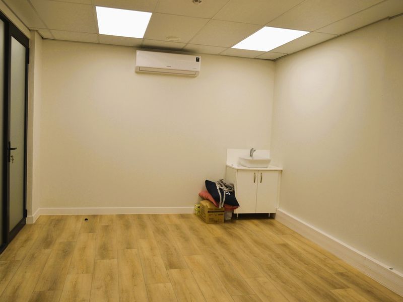 A clean, modern office space featuring wood-look laminate flooring, a suspended ceiling with recessed lighting, a wall-mounted air conditioner, and a small vanity basin.