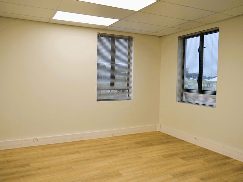 A clean, vacant office space featuring modern laminate flooring, suspended ceilings with recessed lighting, and large windows with fitted venetian blinds.