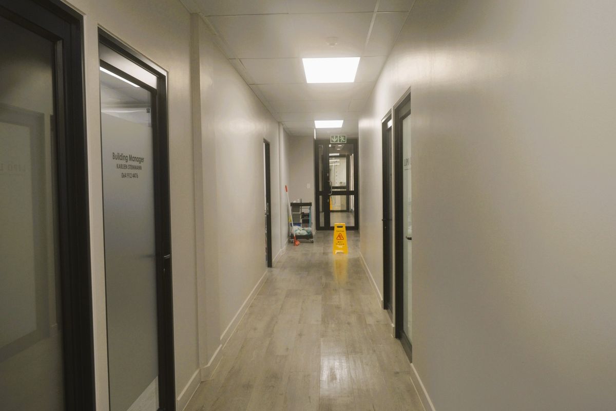 A clean, well-lit commercial office hallway featuring wood-look flooring, suspended ceiling tiles, and multiple glass-paneled office doors.