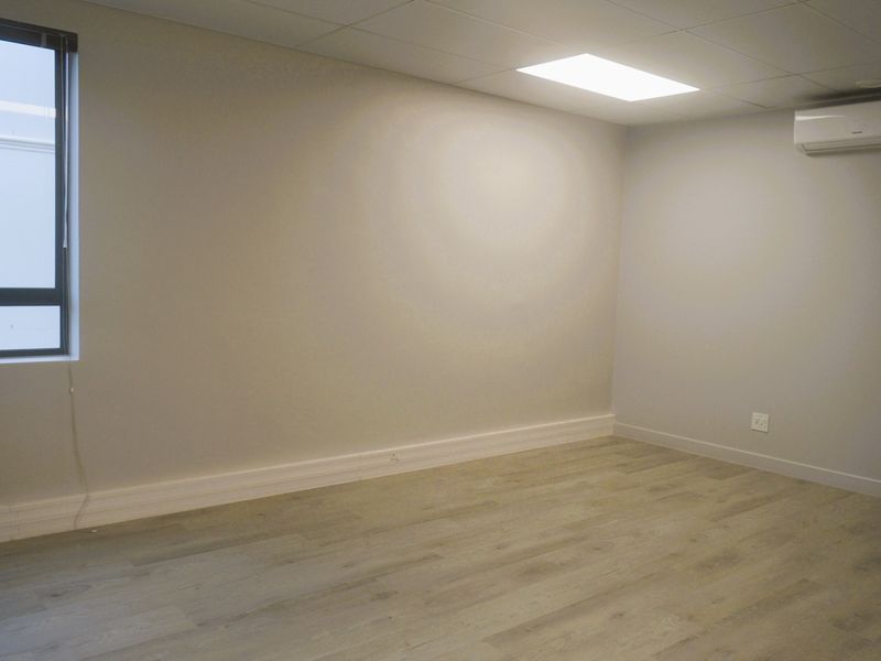 A clean, empty office space featuring modern laminate wood flooring, recessed ceiling lighting, and a wall-mounted split-system air conditioning unit.