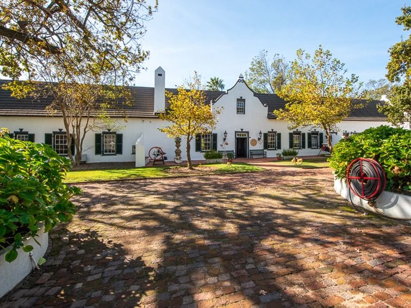A charming Cape Dutch style farmhouse featuring a classic gabled facade, brick-paved driveway, and well-maintained garden beds.