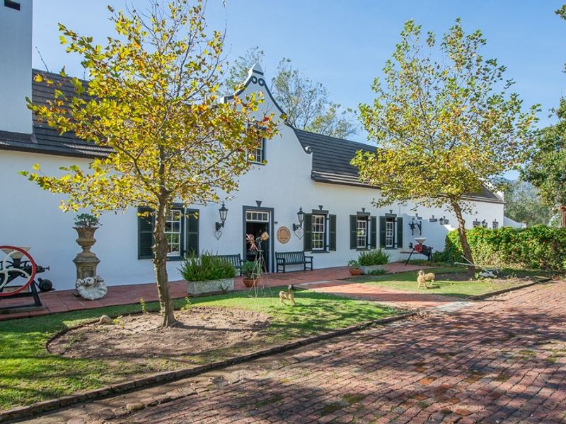A charming Cape Dutch style farmhouse featuring a white facade, traditional gable, dark shutters, and a spacious brick-paved courtyard with mature trees.