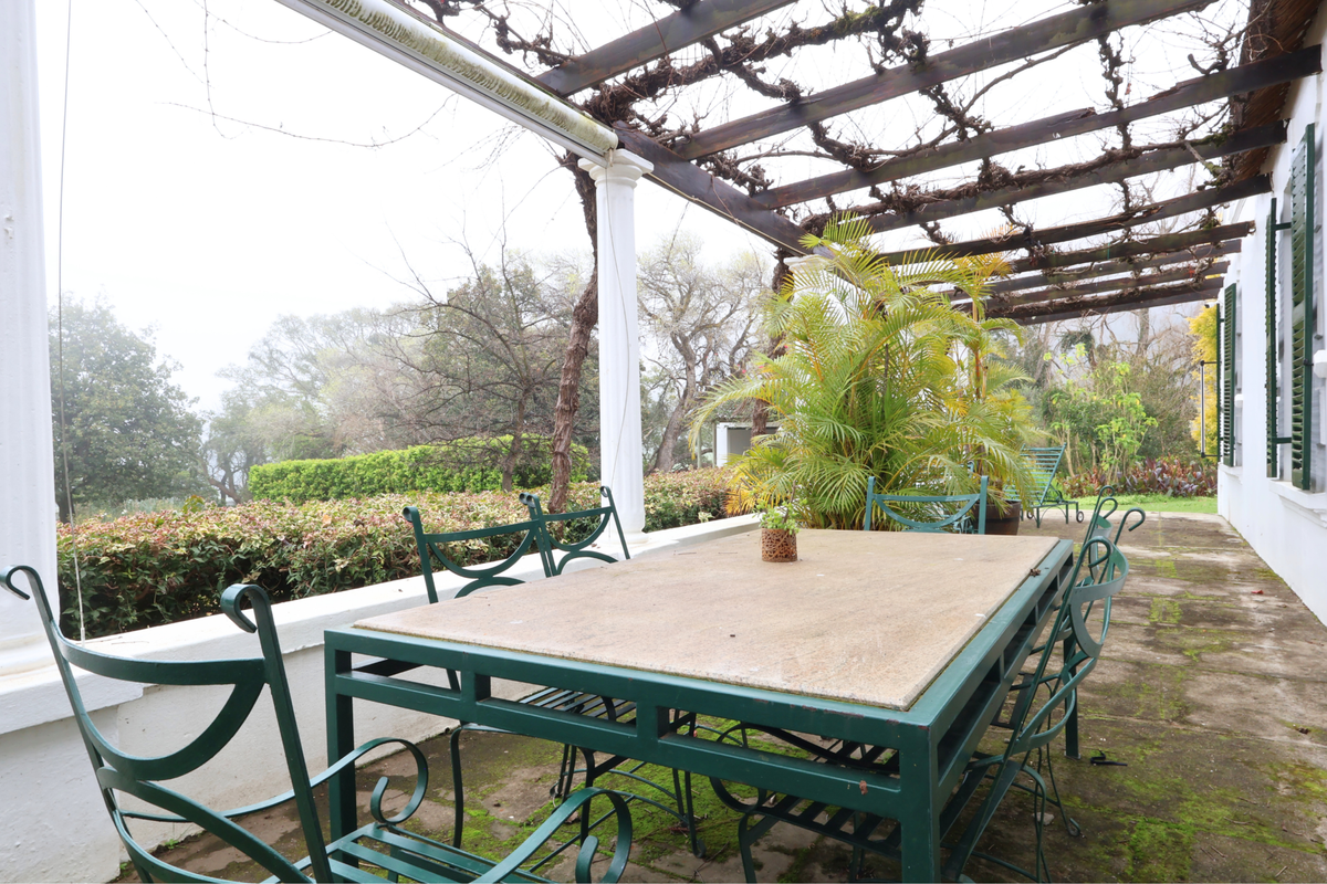 A covered outdoor patio featuring a timber pergola, concrete columns, and paved flooring, overlooking a garden area.