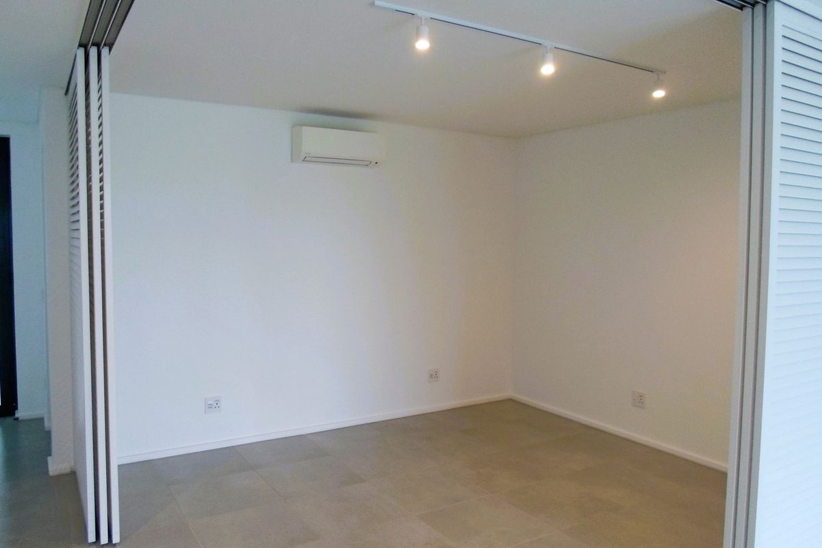 A bright, empty room featuring neutral floor tiles, a wall-mounted air conditioning unit, and modern track lighting, enclosed by folding louvered doors.