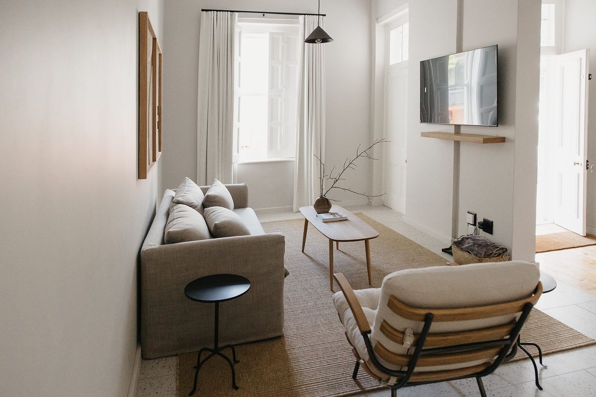 A bright and modern living room featuring terrazzo flooring, a wall-mounted television with a floating shelf, and natural light through shuttered windows.