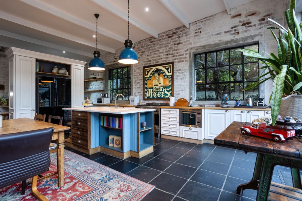 A characterful kitchen featuring exposed brick walls, a central island with blue cabinetry, stone tile flooring, and large metal-framed windows.