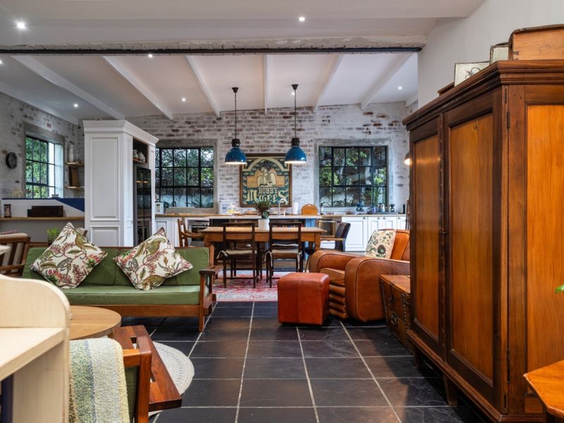 Spacious open-plan living and kitchen area featuring exposed brick walls, timber ceiling beams, stone flooring, and large steel-framed windows providing natural light.