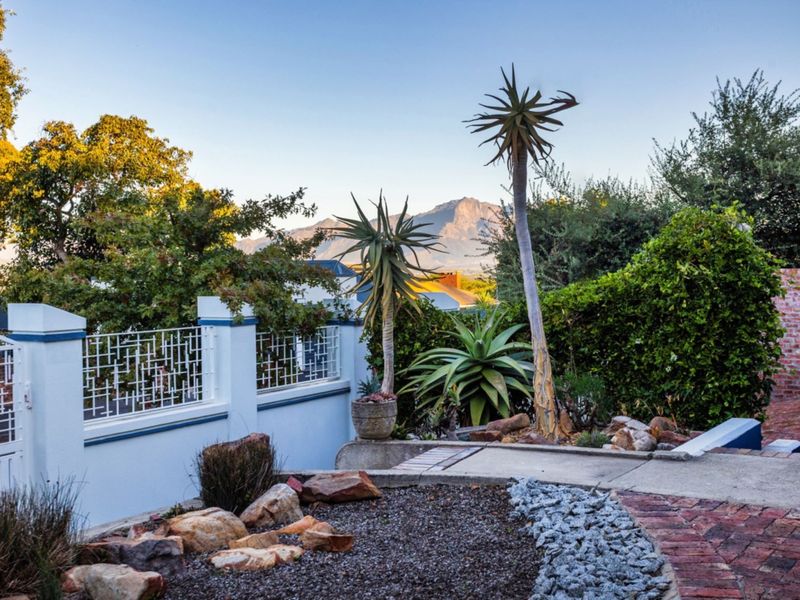 A well-maintained garden area featuring brick and concrete pathways, established landscaping, and a secure boundary wall with mountain views in the background.