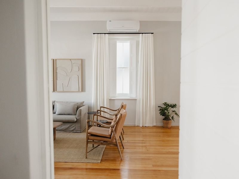 A bright and well-maintained living room featuring polished timber flooring, a sash window with curtains, and a wall-mounted air conditioning unit.