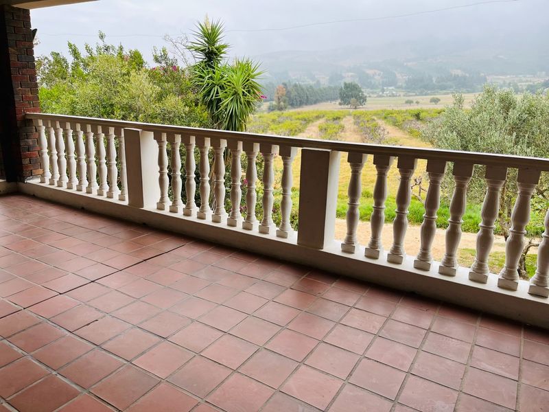 A spacious covered balcony featuring a classic balustrade, terracotta-style tiled flooring, and expansive views over the surrounding landscape.