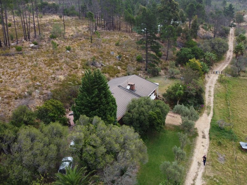 Aerial view of a residential property nestled in a rural landscape with a metal roof, brick chimney, and access via a gravel driveway.