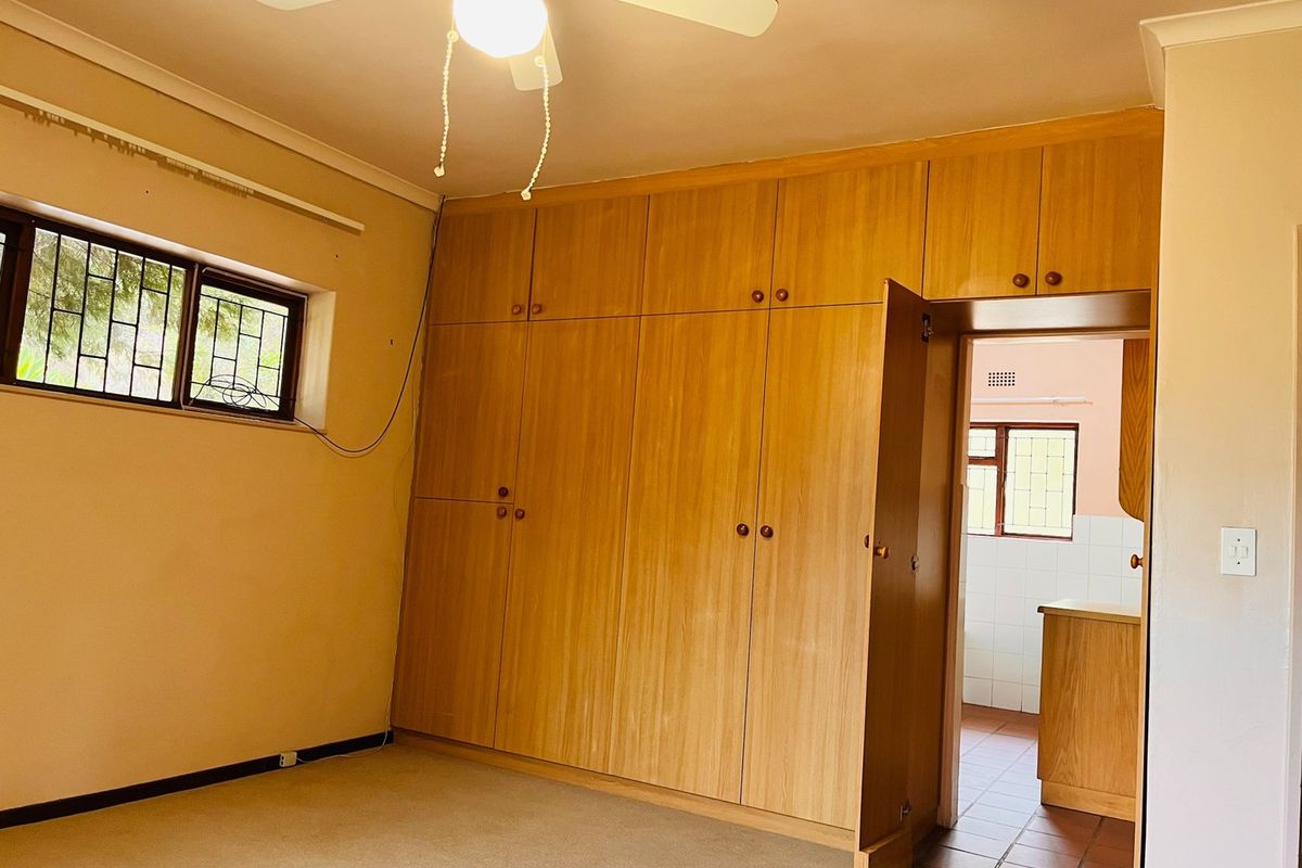 A spacious bedroom featuring extensive built-in wooden cupboards, a ceiling fan, and secure window bars, offering a functional layout with neutral finishes.