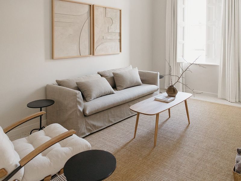 A bright and minimalist living room featuring neutral tones, natural light from a casement window, and clean architectural lines.