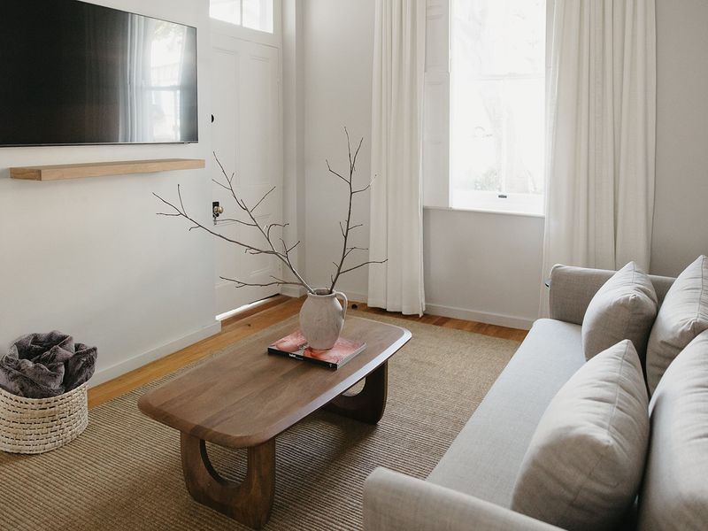 A bright and inviting living room featuring polished hardwood flooring, a minimalist timber floating shelf, and a classic panelled entrance door.