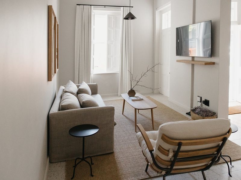 A bright and modern living room featuring terrazzo flooring, a wall-mounted television with a floating shelf, and natural light through shuttered windows.