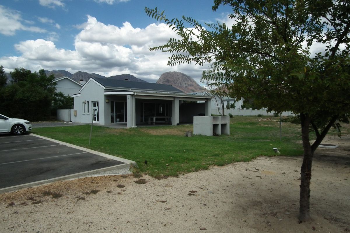 Rear view of the apartment featuring a covered patio, built-in braai area, and adjacent paved parking, set against a scenic mountain backdrop.