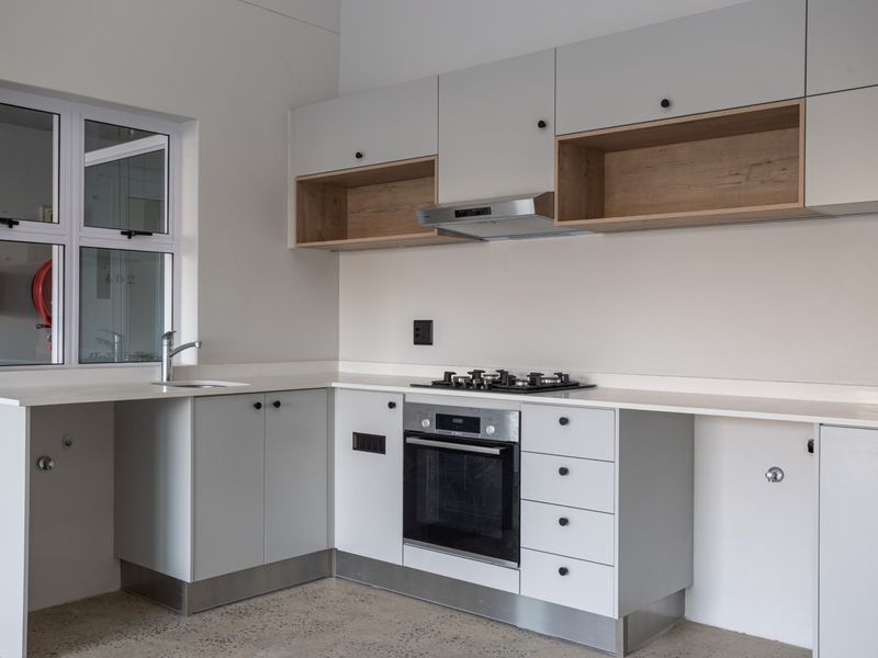 Modern kitchen featuring light grey cabinetry, wood-accented open shelving, stone countertops, integrated gas hob, oven, and extractor fan with polished concrete flooring.