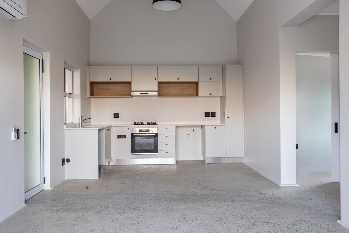 Modern open-plan kitchen featuring sleek white cabinetry, integrated oven and hob, polished concrete flooring, and a high vaulted ceiling in a minimalist space.