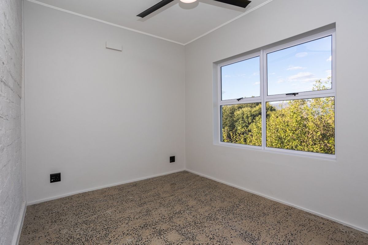 A bright, empty bedroom featuring polished exposed aggregate concrete flooring, a ceiling fan, and a large window with a pleasant green outlook.