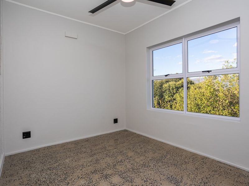 A bright, empty bedroom featuring polished exposed aggregate concrete flooring, a ceiling fan, and a large window with a pleasant green outlook.