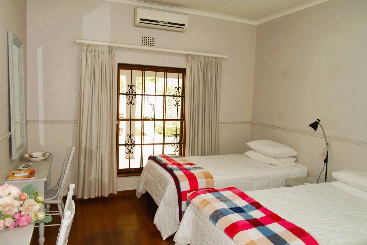 A bedroom featuring wooden flooring, a wall-mounted air conditioning unit, and a window with security bars and curtains.