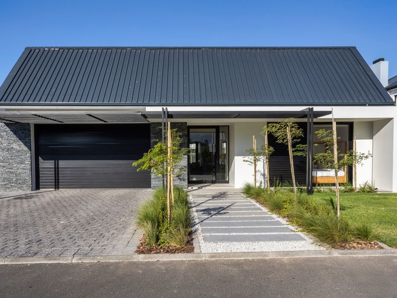Modern single-story house facade featuring a dark metal roof, stone cladding, paved driveway, and a sleek entrance walkway with integrated pergola.