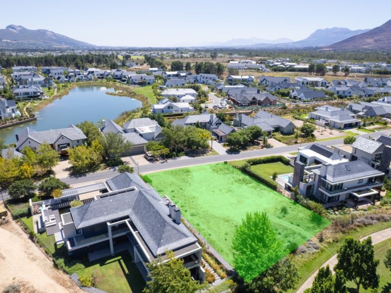 Aerial view of a vacant residential plot situated within a developed estate, featuring surrounding homes, a nearby lake, and scenic mountain views.