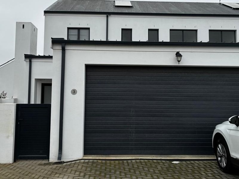 Front elevation of a modern townhouse featuring a dark sectional garage door, paved driveway, and clean white exterior finishes.