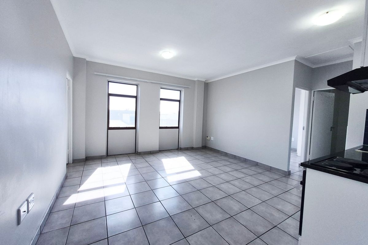 An open-plan living and kitchen area featuring neutral tiled flooring, ample natural light from dual windows, and a functional kitchen layout.
