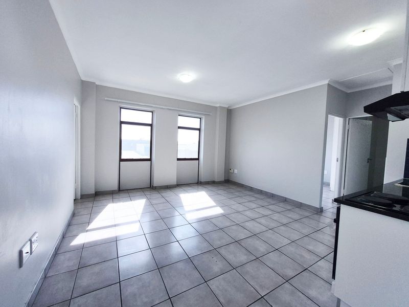An open-plan living and kitchen area featuring neutral tiled flooring, ample natural light from dual windows, and a functional kitchen layout.