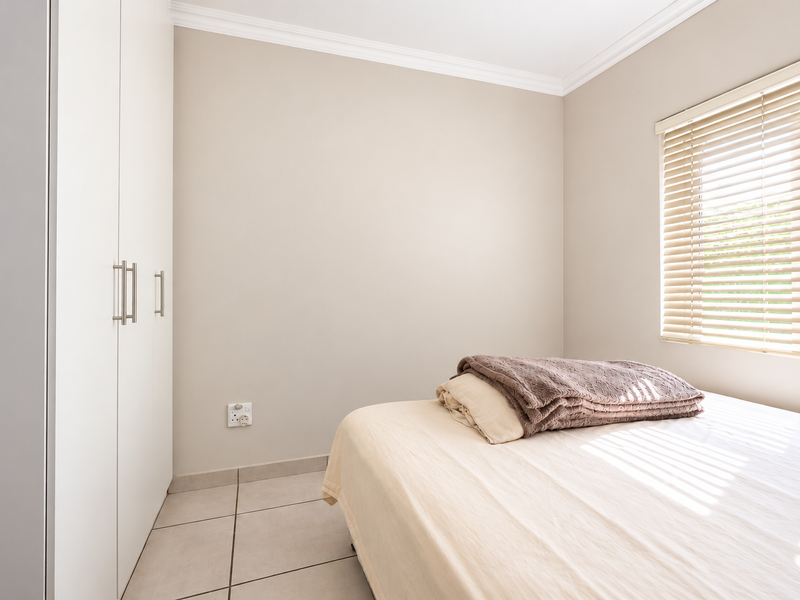 A bright bedroom featuring neutral walls, tiled flooring, built-in cupboards, and venetian blinds allowing natural light.