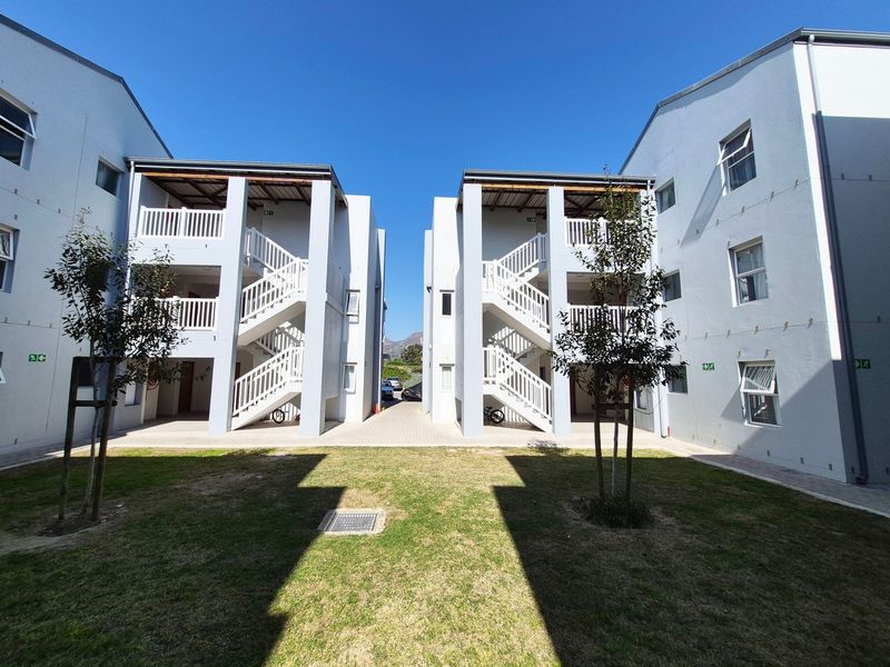 Exterior view of apartment complex buildings featuring central lawn, paved walkways, and external staircases leading to upper-level balconies.