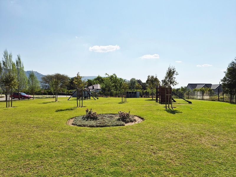 A well-maintained communal garden area featuring a spacious lawn, mature trees, and playground equipment within a secure residential estate.