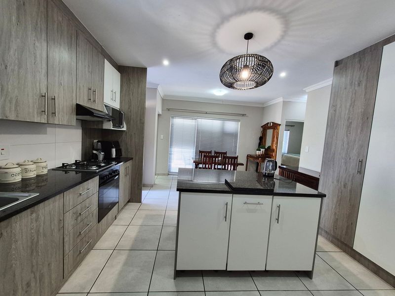 Modern open-plan kitchen featuring wood-grain cabinetry, granite countertops, a central island, and integrated appliances, flowing into a bright dining area with tiled floors.