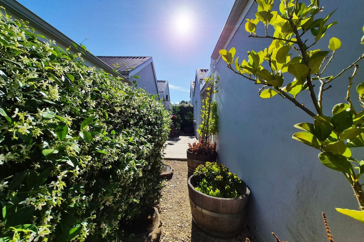 A narrow side garden pathway featuring gravel surfacing, paved patio section, and established perimeter landscaping between townhouse structures.