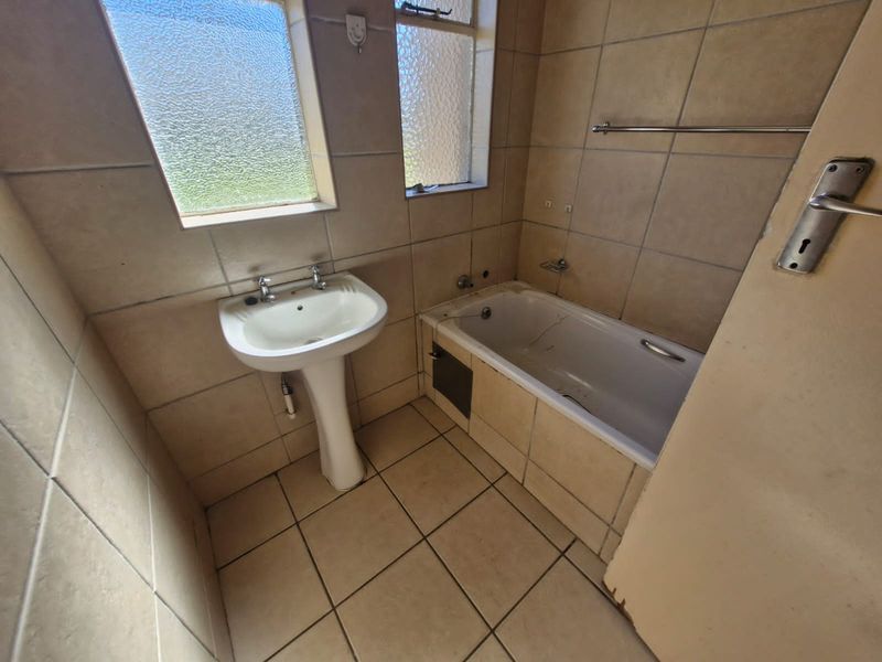 A dated bathroom featuring a pedestal basin, built-in bathtub, and tiled finishes, requiring renovation to address visible wear and damage.