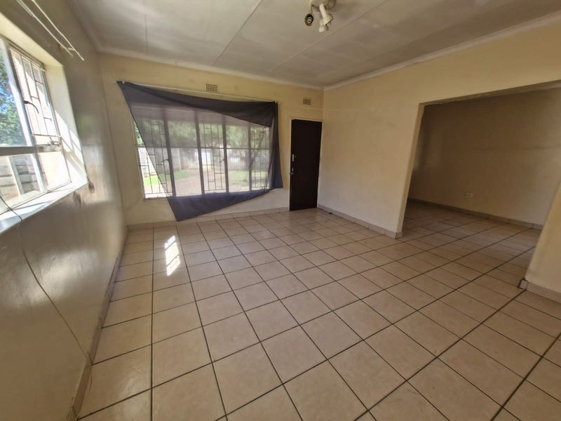 An empty, open-plan living area featuring tiled flooring, security bars on windows, and direct access to an adjacent room.