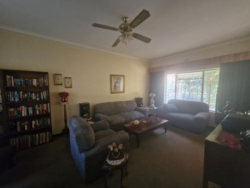 A comfortable living room featuring a ceiling fan, carpeted flooring, and a large window with security bars, offering a traditional and functional living space.