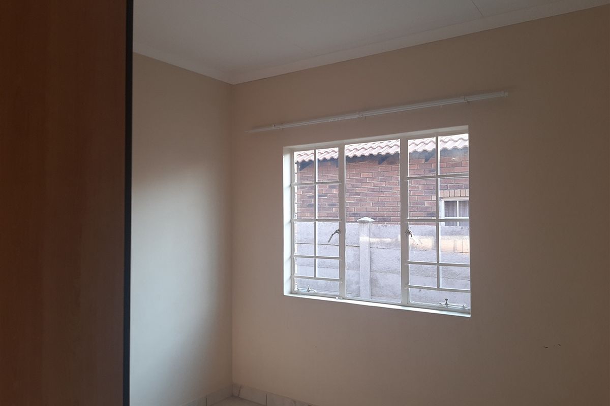 A vacant bedroom featuring neutral wall tones, tiled flooring, and a window with security bars overlooking the exterior brick wall.