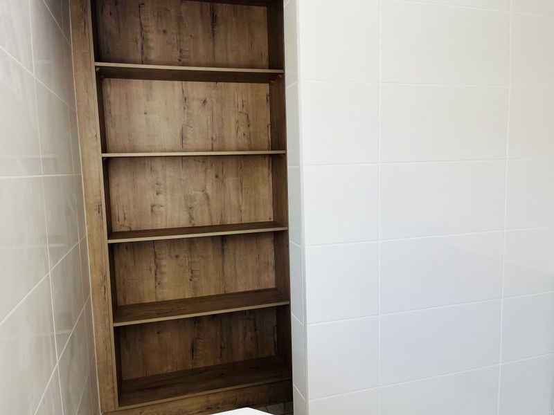 A clean, modern bathroom featuring white ceramic wall tiles and a built-in wood-look shelving unit.