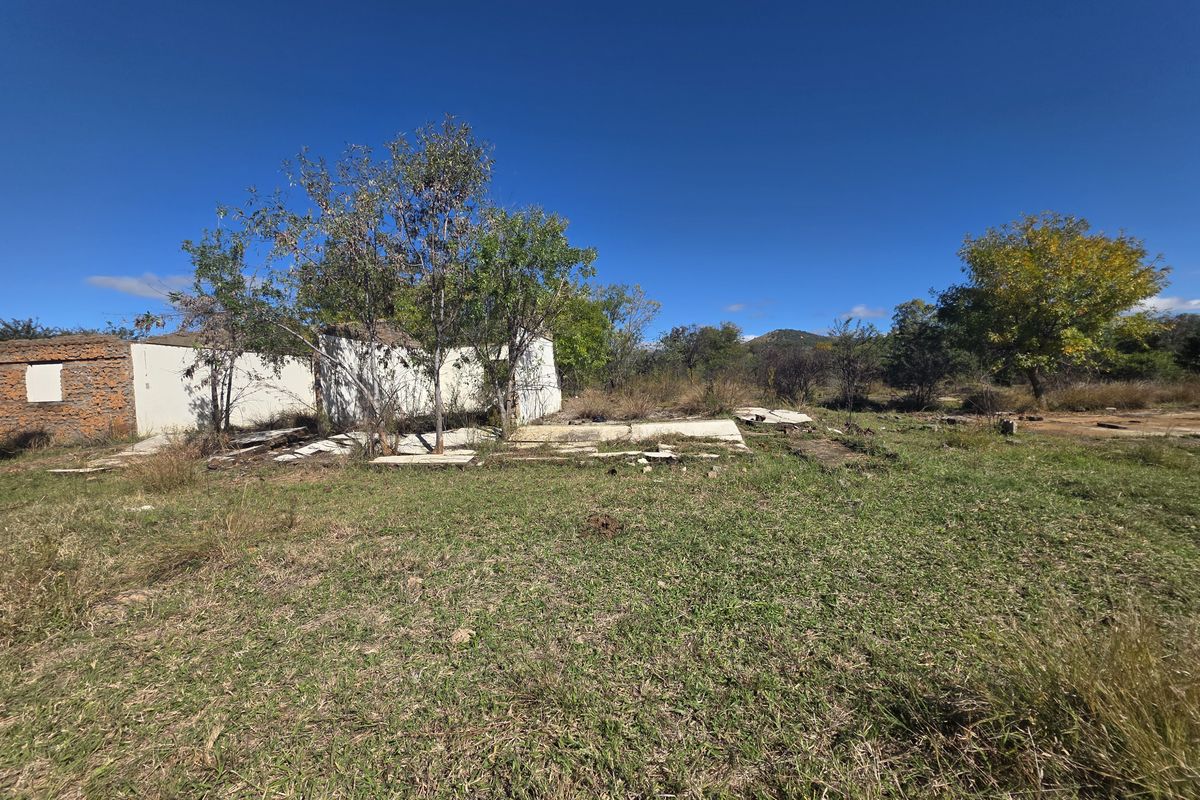 A vacant residential plot featuring the remains of a dilapidated structure and a broken concrete foundation, surrounded by natural landscape and mature trees.