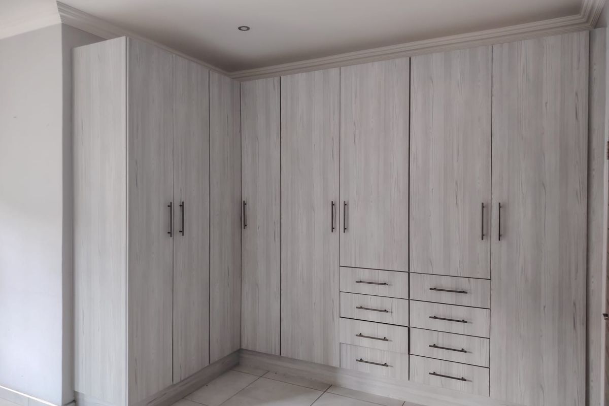 A clean, modern bedroom featuring extensive light wood-grain built-in cupboards with integrated drawers and recessed ceiling downlights.
