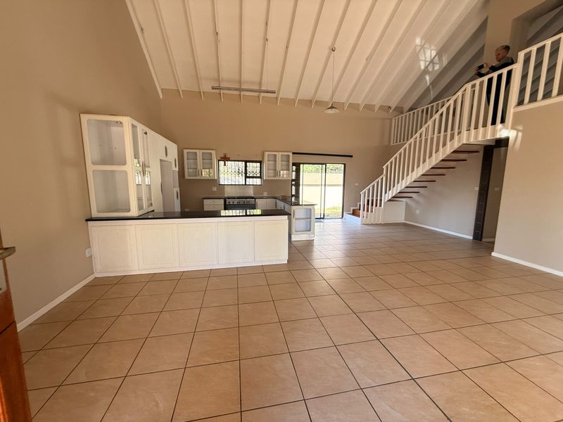 Spacious open-plan living area featuring a kitchen with island, tiled flooring, high exposed beam ceilings, and a staircase leading to a mezzanine level.