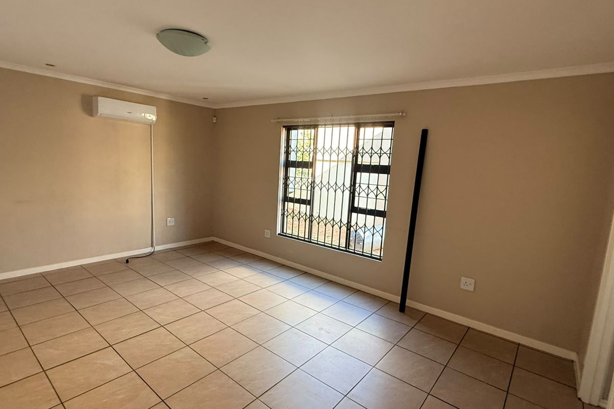 An empty living room featuring tiled flooring, a wall-mounted air conditioning unit, and security bars on the window.