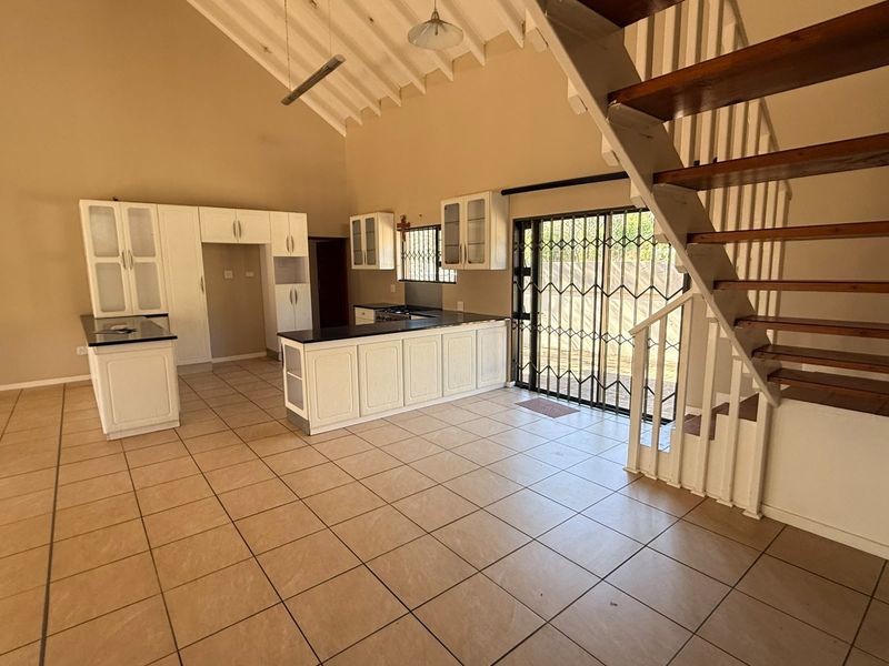 Open-plan kitchen and living area featuring high ceilings with exposed rafters, tiled flooring, built-in cabinetry, and a timber staircase leading to an upper level.