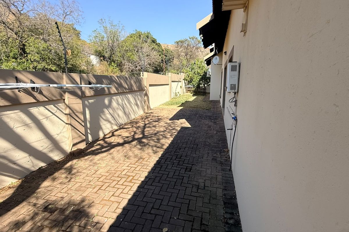 A side exterior view showing a paved walkway, boundary wall with electric fencing, and an external wall-mounted geyser unit.