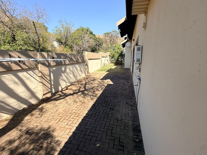 A side exterior view showing a paved walkway, boundary wall with electric fencing, and an external wall-mounted geyser unit.