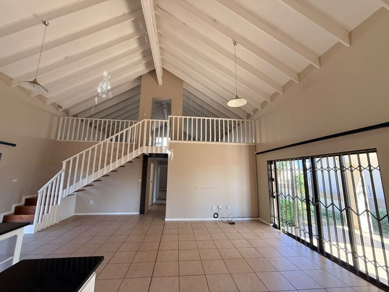 Spacious open-plan living area featuring a high vaulted ceiling with exposed rafters, a mezzanine level, tiled flooring, and large sliding doors with security gates.