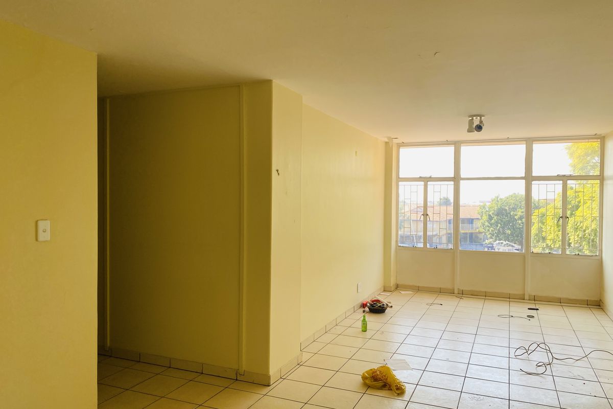 An empty, sunlit living area featuring tiled flooring and large windows with security bars, offering a blank canvas for renovation.