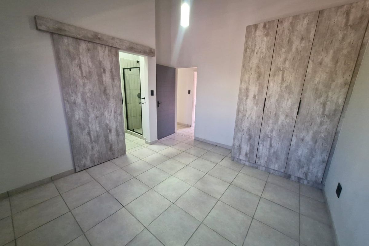 A spacious, empty bedroom featuring modern wood-look built-in cupboards, a sliding barn door leading to an en-suite bathroom, and neutral tiled flooring.